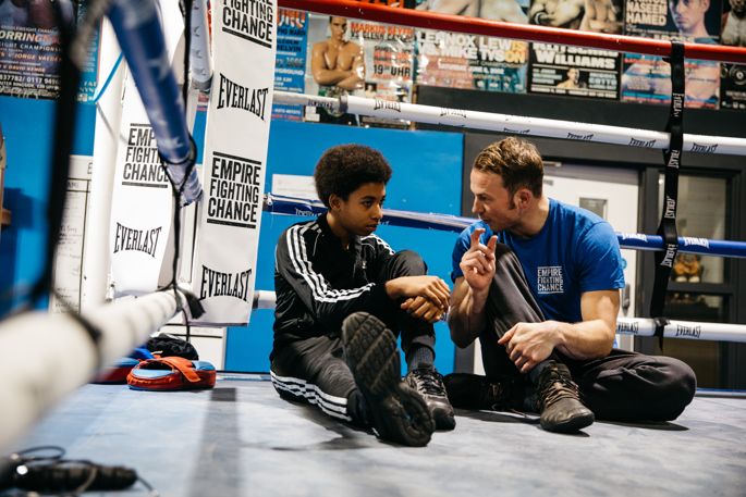 The image shows a young person and their coach chatting together in the boxing ring